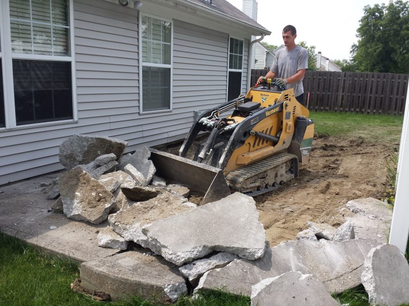 A small skid loader is demolishing and removing a previous patio to prepare for a replacement one. 