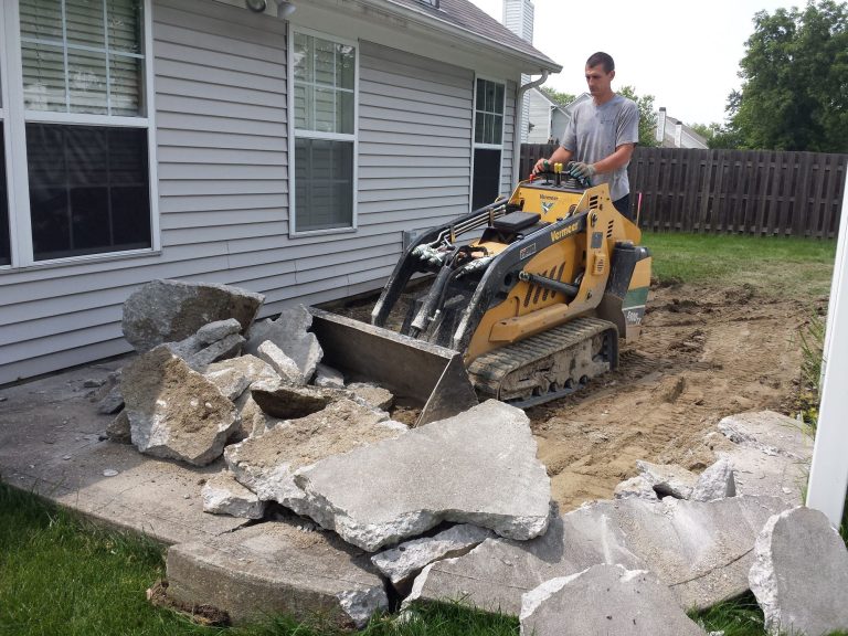 A yellow mini skid steer with a bucket attachment is scooping up broken concrete from a backyard patio. The machine is positioned near a pile of concrete debris, with the bucket on the ground and filled with chunks of concrete. Dust and concrete fragments are visible around the backyard area 