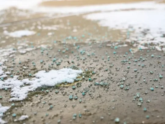 Close-up of a driveway covered with salt to melt snow. The entire surface, including patches of remaining snow, is covered with a layer of salt. The snow is sparse and mostly concentrated in some areas, with the salt visibly spread across the entire driveway