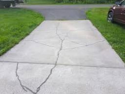 Concrete driveway with large cracks running down the middle. The cracks are deep and wide, creating noticeable gaps and unevenness along the center of the driveway. The surrounding concrete shows signs of wear and aging