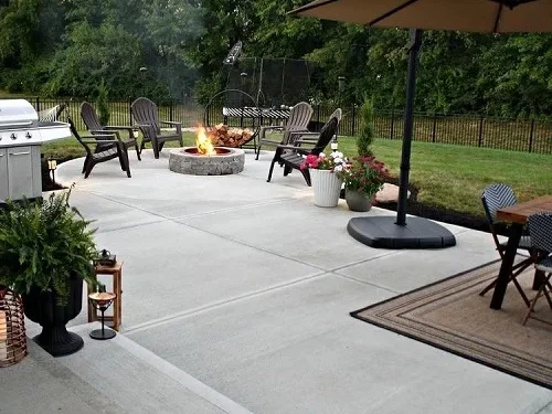 Newly installed outdoor concrete patio with a circular central fire pit surrounded by lounge chairs and leads to a concrete area with a basketball hoop for the whole family to enjoy their backyard