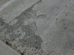 Close-up of a concrete driveway showing damage from weather. The surface features multiple cracks, stains, and discoloration. There are signs of erosion and wear, with some areas appearing chipped or flaked due to exposure to the elements