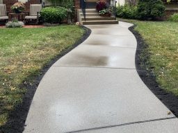 Concrete pathway leading to a home, recently sealed with a glossy finish. The surface is shiny and reflects light, with water droplets visible on it. The pathway appears smooth and well-maintained, highlighting the new sealant and its protective effect.