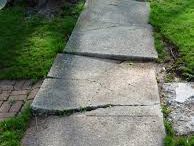 A concrete walkway with multiple cracks and broken sections that need repair or replacement. The surface is uneven with large gaps and chunks missing, and some areas show signs of significant wear and damage