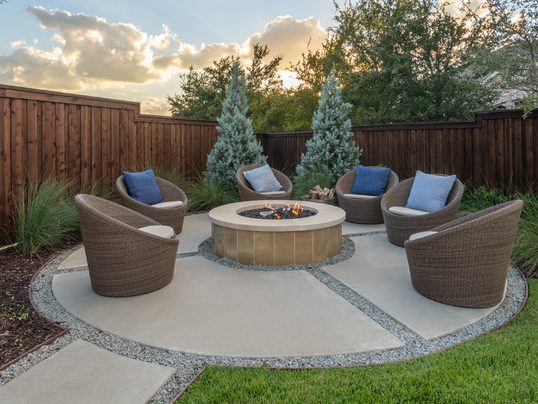 New outdoor concrete patio featuring a round design divided into multiple sections. The patio is outlined by a rock-patterned concrete border, with a round fire pit centrally located. Surrounding the fire pit are modern wicker chairs arranged for hosting, creating a stylish and inviting gathering area