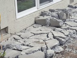 A concrete area next to a home with multiple cracks and broken sections that needs replaced. The surface is uneven with large gaps and chunks of concrete piled up on each other, and some areas show signs of significant wear and damage